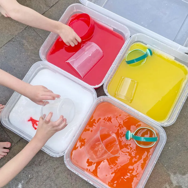 Four bins of colored water sit on a patio, red, orange, yellow, and white. Cups and bowls are in the water and kid hands.