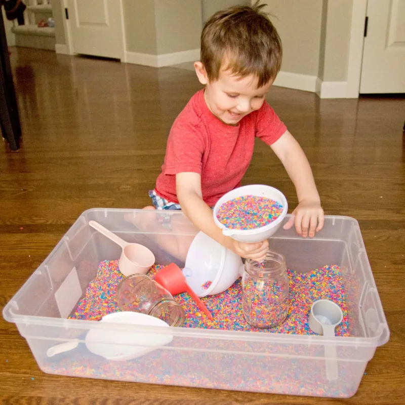 A child smiles while tipping a funnel of rainbow rice into a mason jar. There is a storage container of colorful rice, funnels, and scoops.