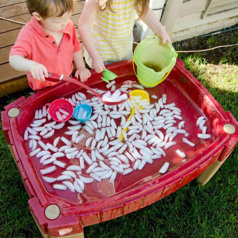 Two children play at a sand table full of ice cubes and water. One is scooping ice while the other holds a bucket.