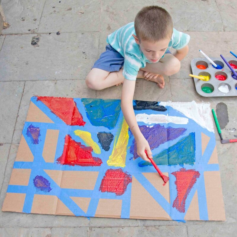 A child leans over a tape resist art project using red paint to fill in a triangle made of blue tape.