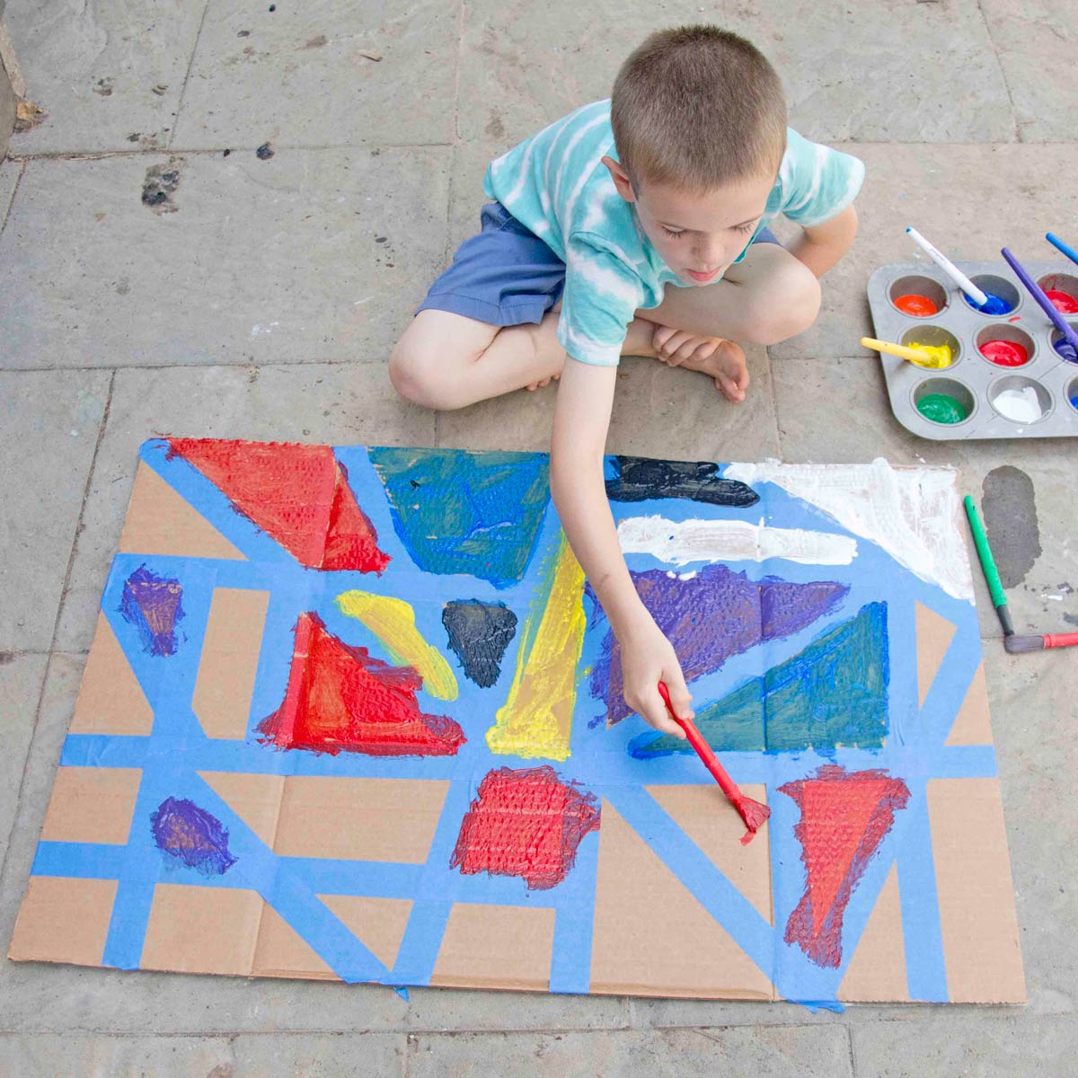 A child leans over a tape resist art project using red paint to fill in a triangle made of blue tape.