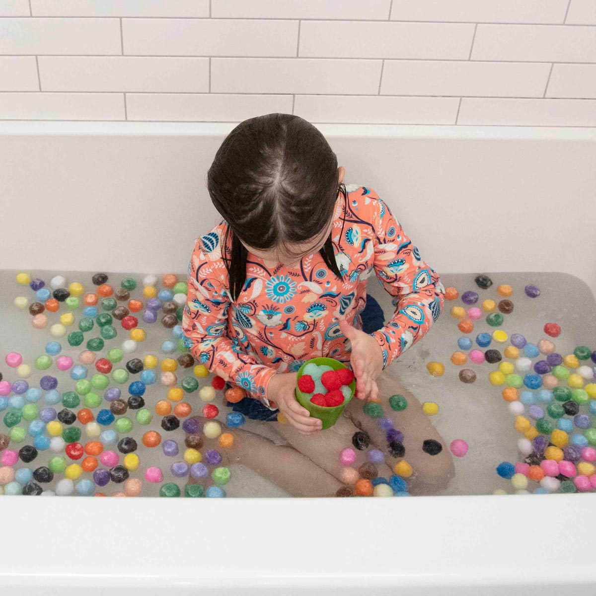 A child sits in a white bath, playing with red and green pom poms in a cup. The bath is filled with multicolored pom poms, making it a vibrant sensory activity for kids.